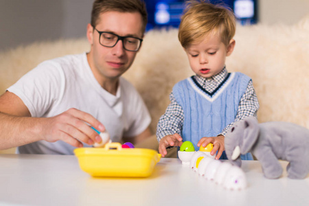 年轻英俊的父亲和他可爱的小儿子坐在桌子旁，在室内玩玩具