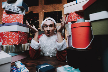 穿着圣诞老人服装带礼品盒的年轻人。 新年前夜。 办公室里的圣诞树。 庆祝新年。 红色服装。 戴红帽子的人。 一堆箱子。 坐着的人