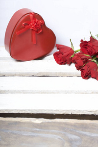 一束猩红玫瑰和一个心形盒子里的礼物。 五朵花躺在一个木箱上。 在白色的背景上。