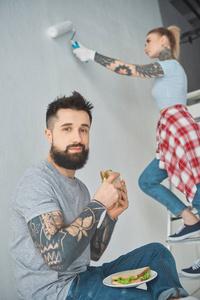 有纹身的男人吃三明治，女朋友在新家画墙
