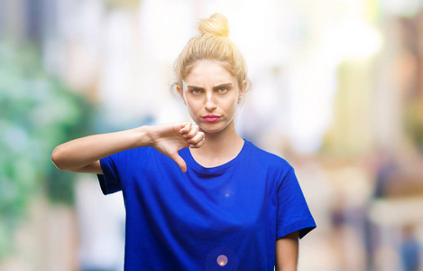 年轻漂亮的金发碧眼的女人穿着蓝色的T恤，在孤立的背景上看起来不高兴和愤怒，表现出拒绝和消极的拇指向下的手势。 不好的表情。