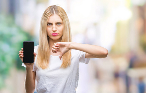 年轻漂亮的金发女人，在孤立的背景上展示智能手机，愤怒的脸，负面的标志，表示不喜欢，拇指向下拒绝概念