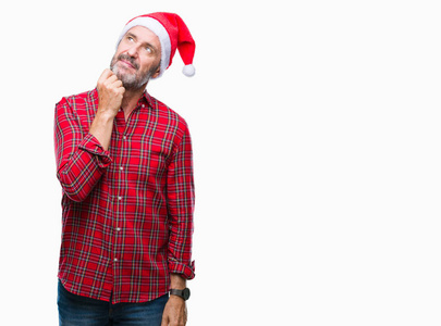 中年老人戴着圣诞帽，戴着孤立的背景，手在下巴上思考问题，沉思的表情。 带着体贴的脸微笑。 怀疑概念。