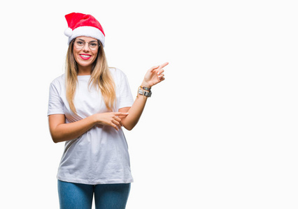 年轻漂亮的女人戴着圣诞帽，在孤立的背景上，脸上挂着一个大大的微笑，用手和手指指向一边，看着相机。