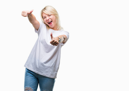 年轻漂亮的金发女人穿着白色T恤，在孤立的背景下，批准用手竖起大拇指做积极的手势，微笑着，为成功而高兴。 看着相机赢家的手势。