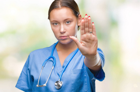 年轻的白种人医生女人穿着医疗制服，在孤立的背景下做停止用手掌唱歌。 警告表情，脸上有负面和严肃的手势。