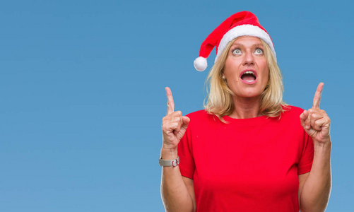 中年金发女人戴着圣诞帽，戴着孤立的背景，惊讶地抬头，用手指和举起的手臂指着。