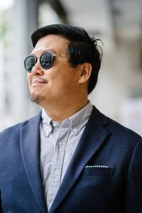 一位中年中国亚裔男子新加坡人的肖像，穿着智能休闲，站在城市里。 他穿着考究，穿着一件海军服，牛津纽扣，衬衫上戴着太阳镜。