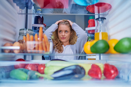 困惑的女人选择吃什么。 手放在头上。 照片是从冰箱里面拍的，里面装满了杂货。