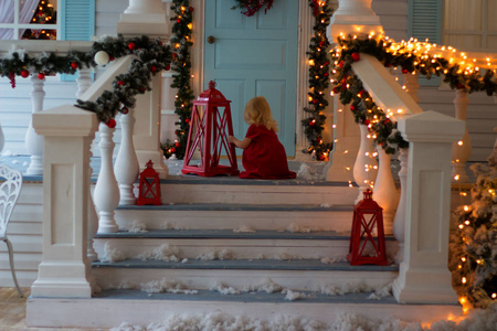 一个穿着红色裙子的小女孩坐在阳台上，楼梯上装饰着灯的花环，看着手电筒。