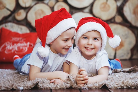 两个戴着圣诞帽的兄弟男孩躺在新年树附近的地毯上，在砍树的背景下大笑。