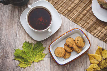 在秋叶背景下，一个古老的茶壶放在稻草床上，上面有一杯热茶，一片李子海绵蛋糕和巧克力饼干。