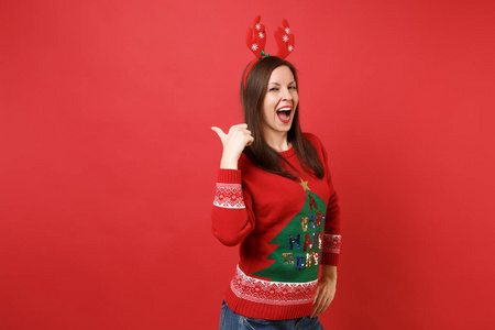 美丽的年轻圣诞老人女孩在有趣的装饰鹿角在头上，保持嘴宽，张开，拇指在后面，孤立的红色背景。 新年快乐2019庆祝节日派对概念