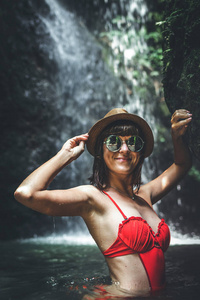 年轻的妇女游客在红色泳装和草帽摆在深丛林与瀑布。真正的冒险概念。巴厘岛