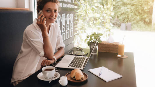 年轻的女学生带着笔记本电脑坐在咖啡馆里，用笔记本电脑做早餐。 幸福的女人自由职业者在咖啡馆用手机说话。