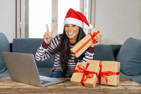 穿着红毛衣和圣诞老人帽的女人在网上选择和购买圣诞礼物，用笔记本电脑坐在家里客厅的沙发上，兴奋地准备好迎接圣诞节