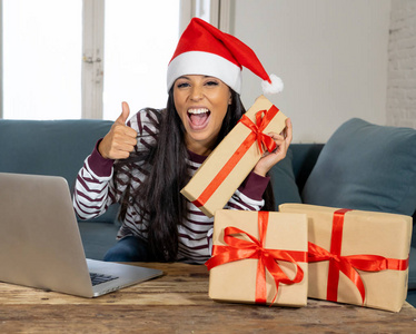 穿着红毛衣和圣诞老人帽的女人在网上选择和购买圣诞礼物，用笔记本电脑坐在家里客厅的沙发上，兴奋地准备好迎接圣诞节