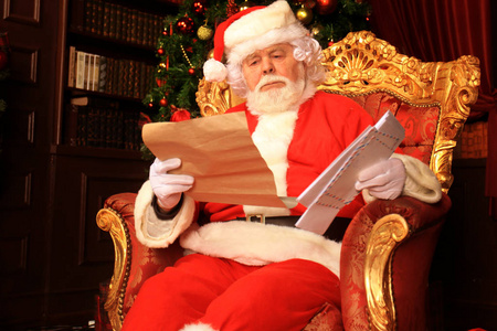 幸福的圣诞老人的肖像坐在他的房间在家里附近的圣诞树和阅读圣诞信函或愿望列表