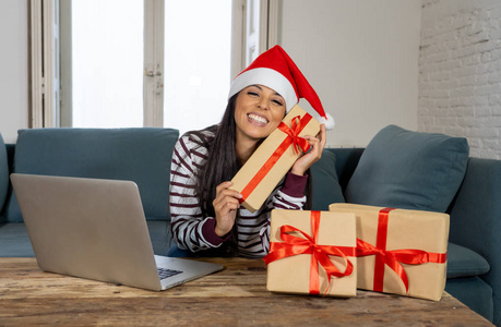 穿着红毛衣和圣诞老人帽的女士在网上选择和购买圣诞礼物，使用笔记本电脑坐在家里客厅的沙发上，兴奋地准备好圣诞礼物