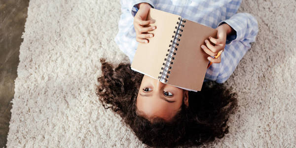 可爱的非洲裔美国儿童，穿着睡衣躺在地毯上，用房间里的拷贝簿遮住脸