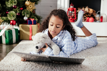 可爱的非洲裔美国儿童穿着睡衣，带着泰迪熊读书在地板上在家圣诞概念