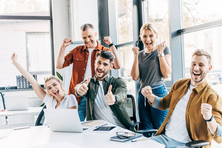 一群快乐的成功企业家庆祝胜利，在办公室竖起大拇指，看着镜头