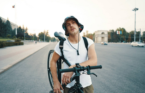 城市中一个金发白人的肖像，肩膀上有一辆经典的自行车，站在城市的一条路上看着镜头
