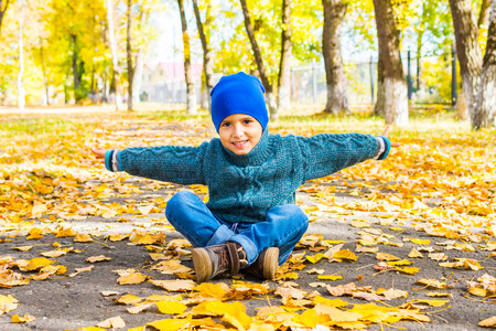 微笑的男孩戴着帽子和毛衣，在秋天坐在公园的跑道上。到处都是黄色的叶子
