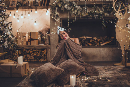 年轻美丽的亚洲微笑的女人穿着睡衣坐在家中靠近壁炉的圣诞树舒适的内部。室内有圣诞装饰。