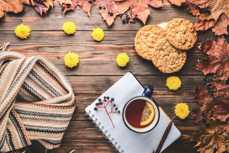 秋季时尚季节性概念黄色温暖柔软毛衣，斯堪的纳维亚针织围巾杯，热红茶，咖啡，落叶，木桌上，笔记本笔。 平躺式顶部复古风格