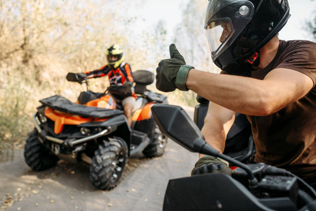 戴着头盔的ATV骑手向他的搭档竖起大拇指，在森林里进行越野旅行。 骑四轮自行车极限运动和旅行四轮自行车冒险