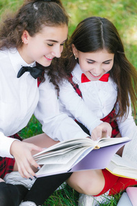 两个有趣的女学生坐在草地上看书。 女孩，女朋友，姐妹都是在大自然中学习的。