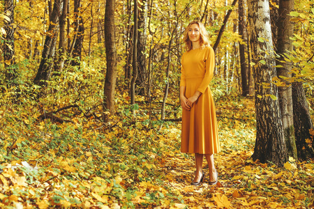 年轻漂亮的女孩穿着黄色的长裙，带着落叶在秋天的公园散步。