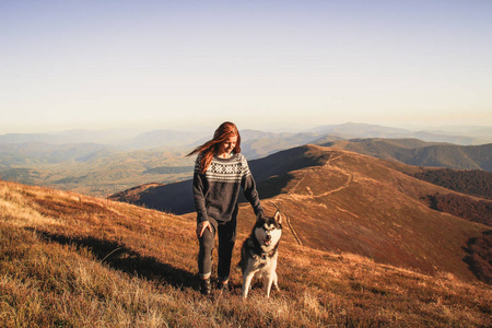红色头发的女孩穿着蓝色毛衣，带装饰品，玩哈士奇。 秋天的乌克兰喀尔巴阡山脉。 温暖的彩色绿叶。森林和山脉的顶部景色。 和狗一起旅