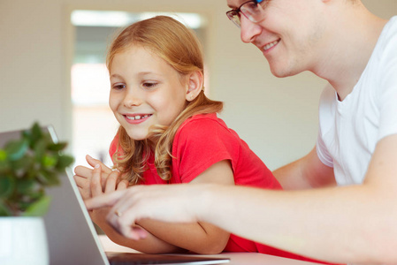 快乐的年轻父亲和他漂亮的小女儿在现代家庭工作时玩笔记本电脑