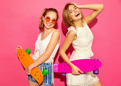 两个年轻时尚微笑的金发女人带着廉价的滑板。 模特在夏季时髦的白色衣服摆在粉红色墙壁附近的工作室太阳镜。 女性