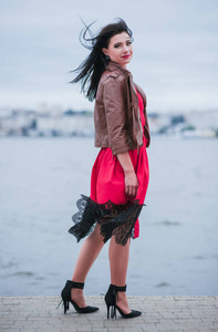 迷人的快乐女人站在湖边，风吹着她的头发城市背景。 穿红色裙子的深色模特在户外摆姿势的垂直镜头