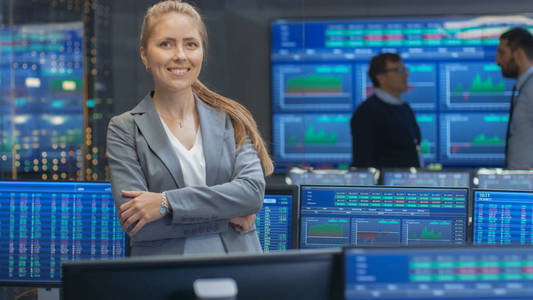 成功的女性股票交易员在镜头前交叉手臂和微笑。在后台繁忙的证券交易办公室与交易员，经纪人和交易商出售和购买债券。显示显示数字.