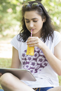 美丽的年轻女孩青少年坐在草地上使用数字平板电脑，喝她最喜欢的橙汁。 4.她喜欢晴朗的夏日