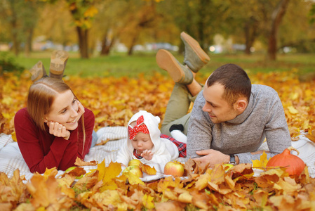 恋爱中的夫妇躺在公园里的秋天落叶上，躺在地毯上，享受着美丽的秋日。 快乐快乐的年轻家庭父亲母亲和小女孩在户外玩得很开心。