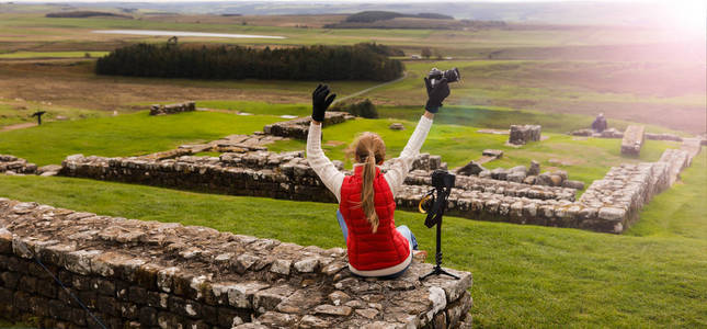 摄影师旅行者坐在相机附近，举起双手。 多云的秋天天气。 美丽的大自然。 背景上的古代石头遗址。