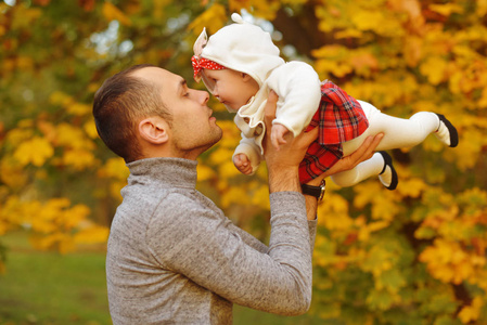父亲和女儿在公园里秋天落叶，享受着美丽的秋日。 快乐快乐的年轻家庭父亲小宝贝女孩在户外玩得很开心。 父母亲吻飞翔的孩子