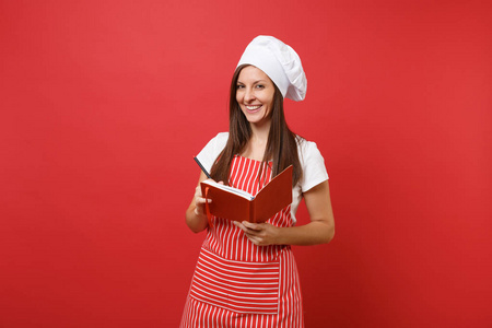 家庭主妇女厨师或面包师穿条纹围裙，白色T恤，烤厨师帽子隔离在红墙背景上。 微笑的女人拿着记事本食谱烹饪书和钢笔。 模拟复制空间概