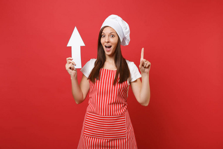 家庭主妇女厨师或面包师穿条纹围裙，白色t恤，烤厨师帽子隔离在红墙背景。微笑幸福的管家女人手里拿着箭。复制空间概念