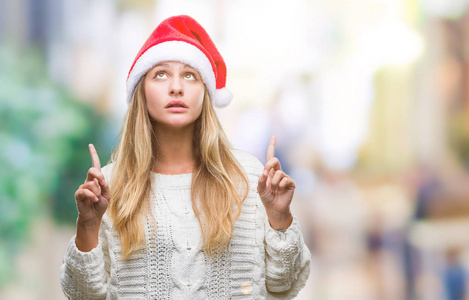 年轻漂亮的金发女人戴着圣诞帽，戴着孤立的背景，惊讶地抬头，用手指和举起的手臂指着。