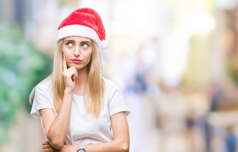 年轻漂亮的金发女人圣诞帽在孤立的背景上，手在下巴上思考问题，沉思的表情。 带着体贴的脸微笑。 怀疑概念。
