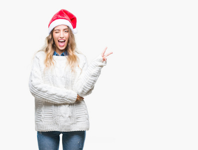 美丽的年轻金发女人戴着圣诞帽，在孤立的背景上微笑，快乐的脸对着镜头眨眼，做胜利标志。 二号。