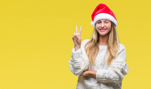 年轻漂亮的金发女郎戴着圣诞帽，戴着孤立的背景，微笑着，快乐的脸对着镜头眨着眼睛，做着胜利的标志。第二名。
