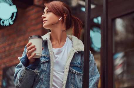 一个穿着牛仔外套的美丽女孩拿着杯子, 在咖啡馆附近的外卖咖啡