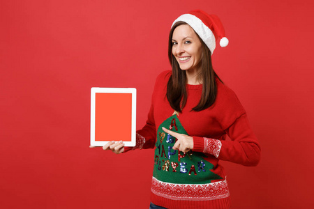 微笑的年轻圣诞老人女孩指着食指在平板电脑电脑上，空白的空屏幕隔离在红墙背景上。 新年快乐，2019年庆祝节日聚会的理念。 模拟复